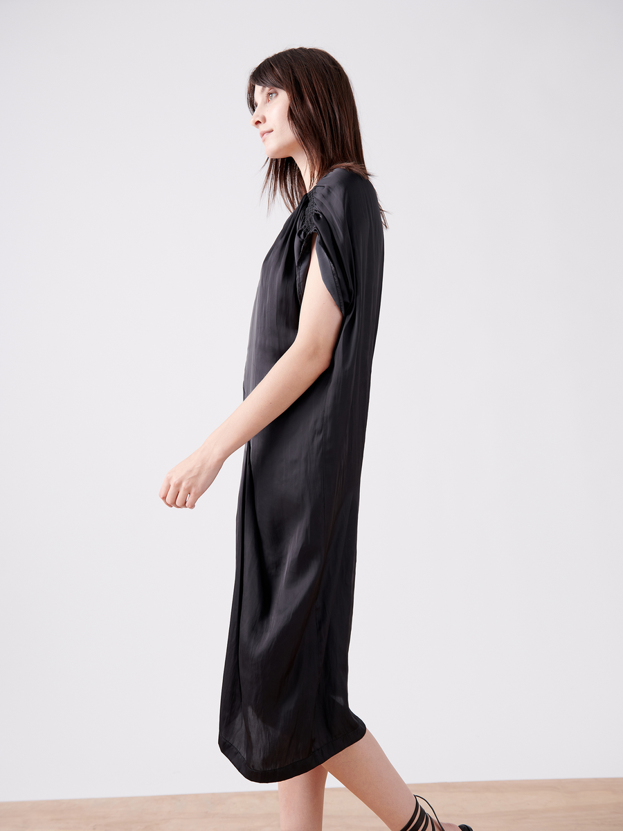 A person with long brown hair walks on a light wooden floor, wearing the Ruched Fold Dress - Final Sale in shiny black by Zero + Maria Cornejo. The asymmetrical folded front contrasts against a minimalist white background.