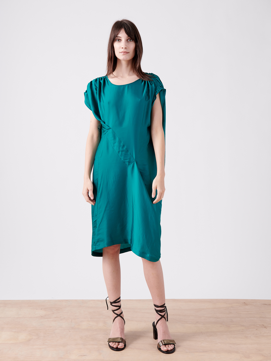 A woman poses in a teal Ruched Fold Dress by Zero + Maria Cornejo, featuring an asymmetrical front, paired with black strappy sandals against a plain white background, looking directly at the camera.