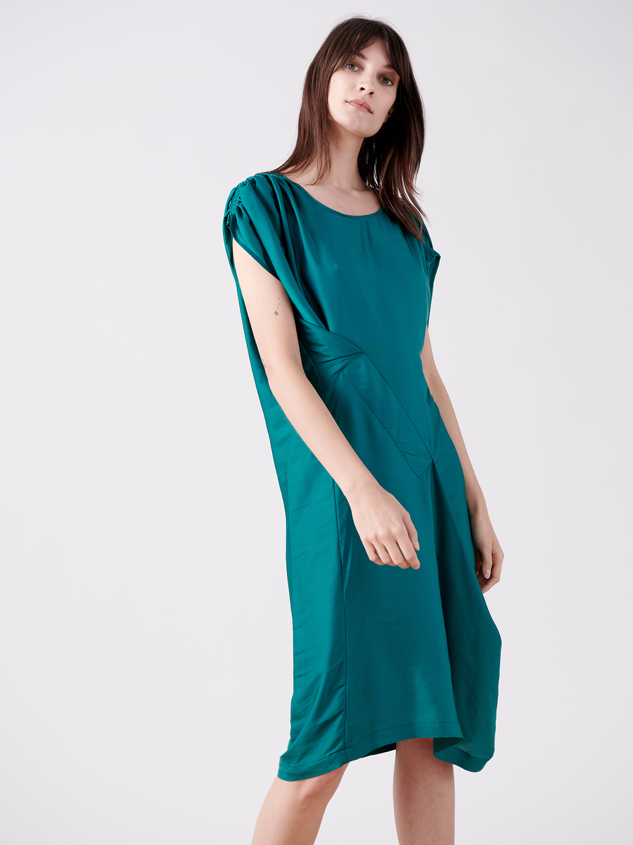 A person with long, straight hair models Zero + Maria Cornejo's Ruched Fold Dress - Final Sale, a teal washed georgette dress featuring gathered fabric at the shoulders and an asymmetrical hem, against a plain white background.