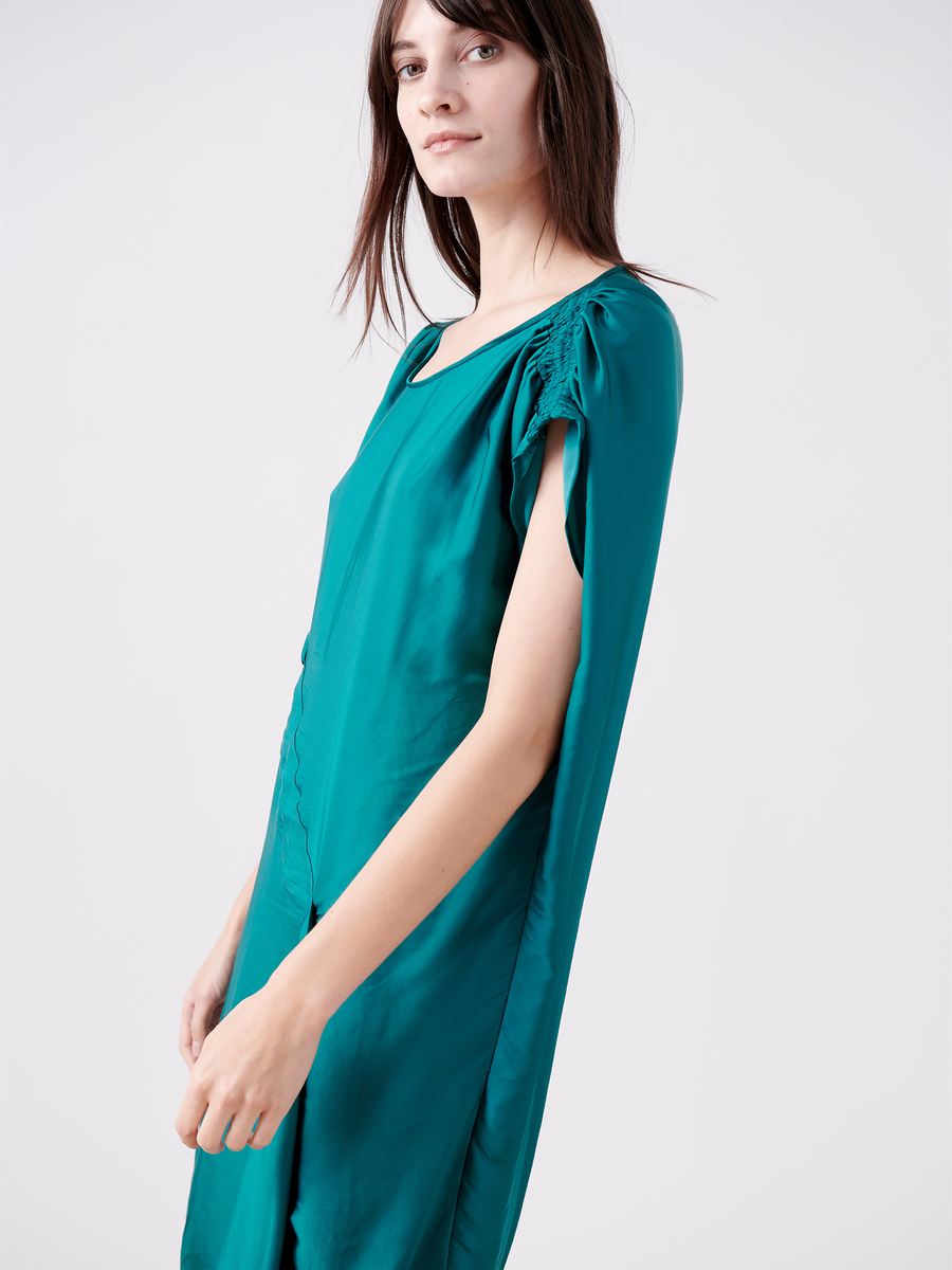 A woman wears the Ruched Fold Dress by Zero + Maria Cornejo, showcasing its teal washed georgette fabric with cape sleeves against a plain background. Her loose long brown hair complements the dress's asymmetrical folded front and gathered shoulder details for added elegance.