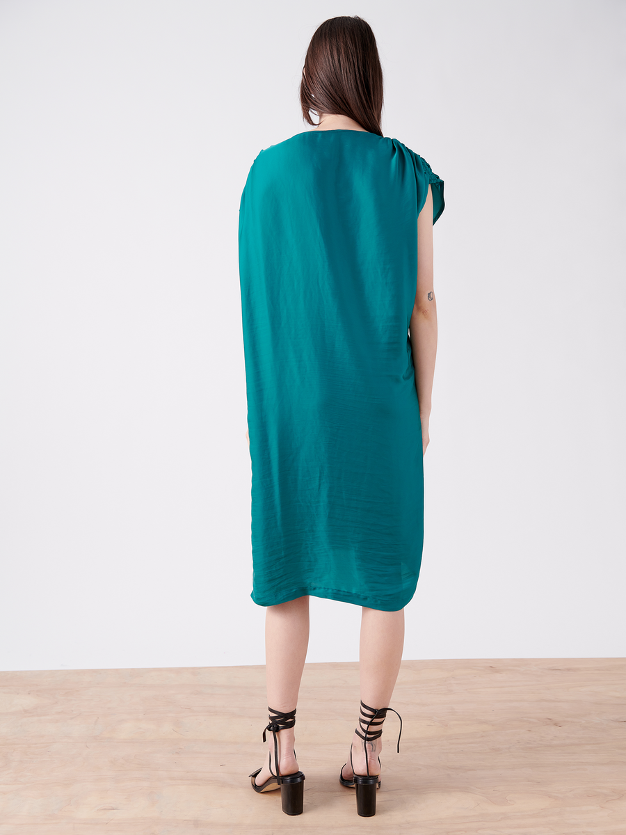 A person with long hair stands facing away, wearing a sleeveless Ruched Fold Dress by Zero + Maria Cornejo in teal washed georgette and black strappy high heels. The dress has an asymmetrical folded front draping loosely, while their arms hang at their sides against a plain background.