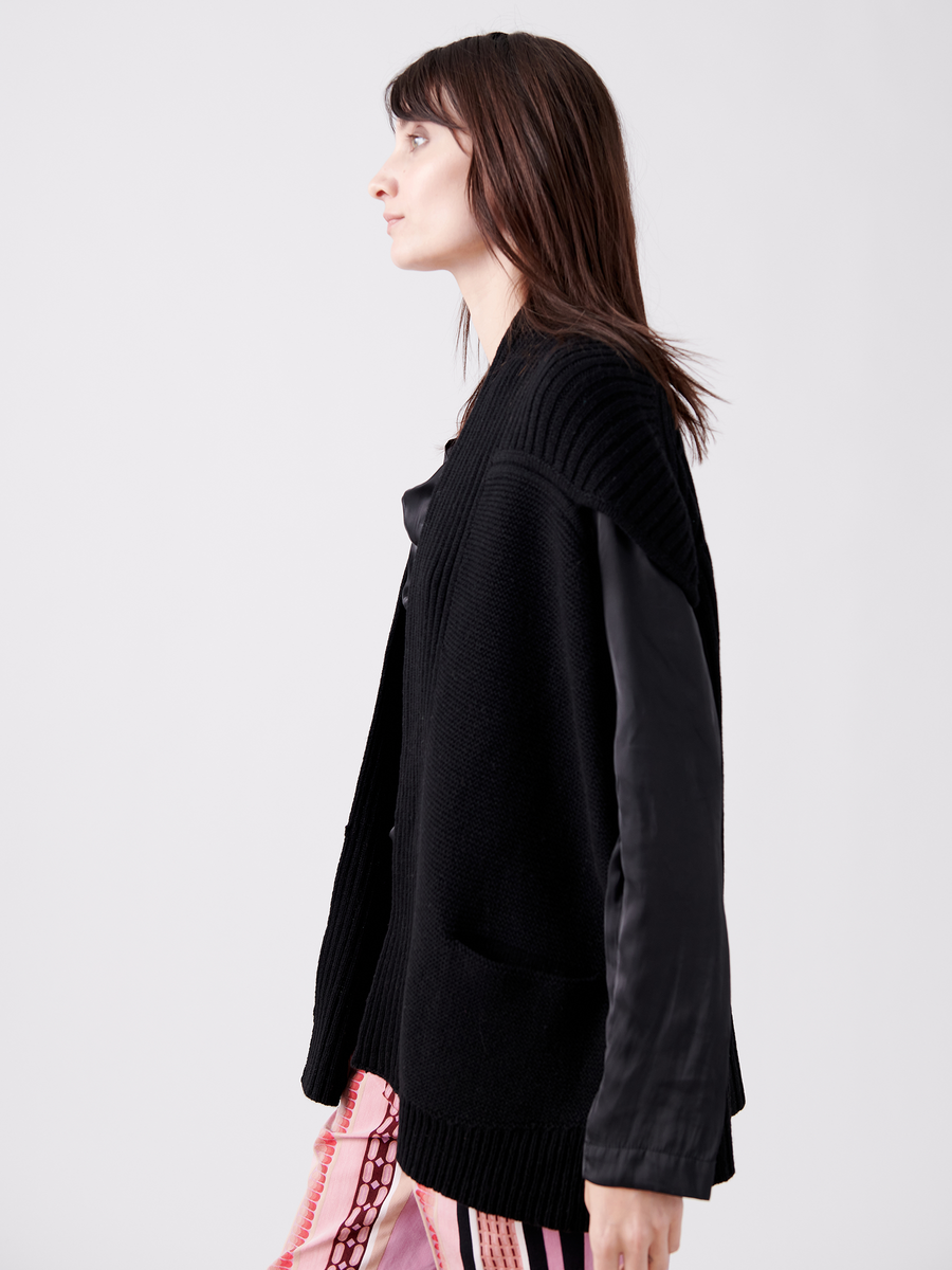 A person with long brown hair wears the Nian Shrug by Zero + Maria Cornejo, embracing sustainable fashion in a black oversized recycled cashmere cardigan and patterned skirt against a plain white background.