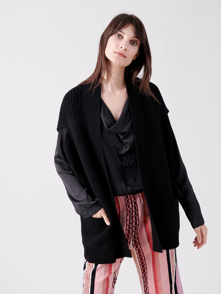 A woman with long brown hair wears the Zero + Maria Cornejo Nian Shrug—a black open cardigan made from recycled cashmere—over a black top and pink striped pants. She stands against a plain white background, embracing sustainable fashion as she looks into the camera.