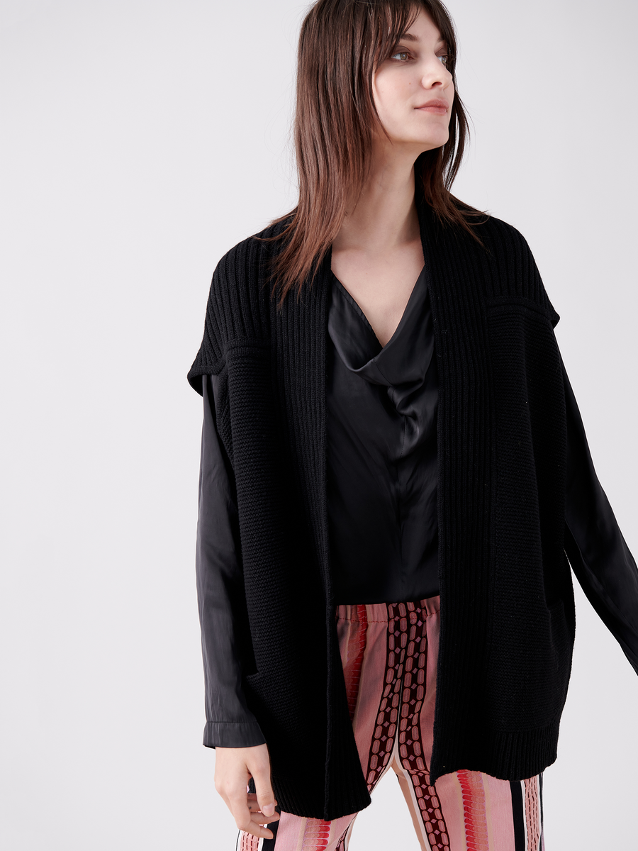 A woman models the Nian Shrug, a black knit cardigan by Zero + Maria Cornejo, crafted from eco-friendly yarn. She pairs it with a black blouse and pink and black patterned pants against a plain white background, slightly angled to the side.