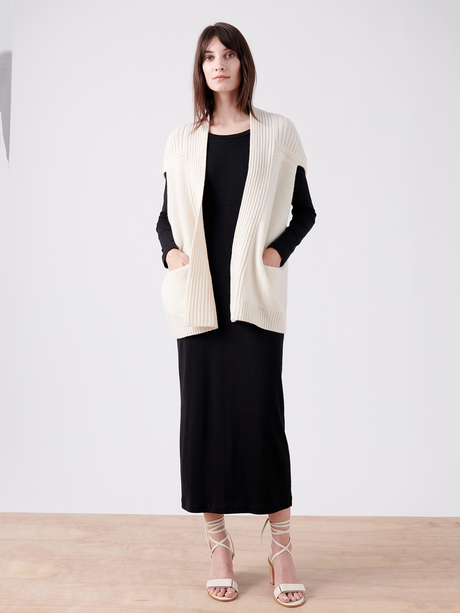 A woman with long dark hair stands against a plain background in a Nian Shrug by Zero + Maria Cornejo, crafted from eco-friendly yarn. She wears a long black dress and white strappy sandals, hands in the pockets of the cream-colored open-front cardigan, embracing sustainable fashion.