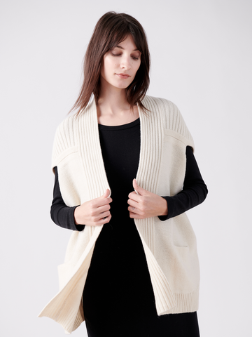 A person with long dark hair wears the Nian Shrug - Final Sale from Zero + Maria Cornejo, a black long-sleeve dress, and a cream-colored ribbed cardigan made from recycled cashmere. They pose against a white background, elegantly holding the cardigan's edges to highlight their sustainable fashion choice.