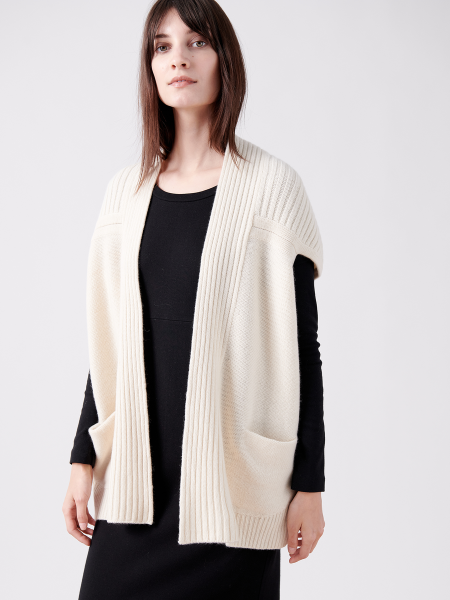 A woman effortlessly embodies sustainable fashion, wearing the Nian Shrug by Zero + Maria Cornejo—a cream-colored, sleeveless knit cardigan made from eco-friendly yarn, featuring wide ribbed edges and front pockets—set against a plain white background.