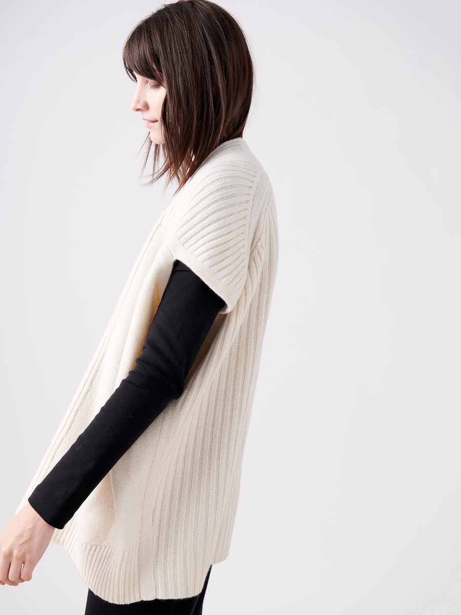 Wearing the Nian Shrug from Zero + Maria Cornejo, a person with shoulder-length hair showcases a cream ribbed knit cardigan made from recycled cashmere over a black long-sleeve shirt. They stand in profile against a light background, epitomizing sustainable fashion.