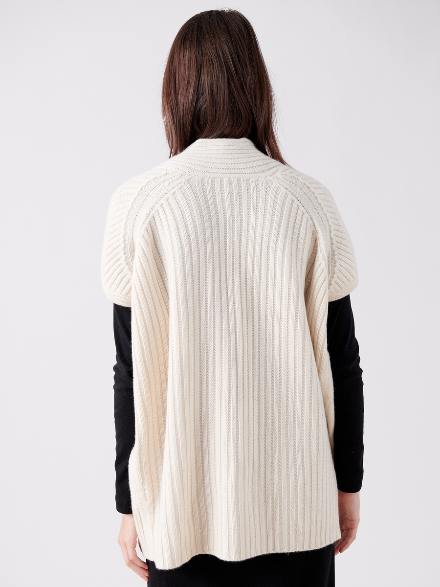 Rear view of a person with long brown hair wearing the Nian Shrug by Zero + Maria Cornejo. This cream-colored, ribbed knit cape, made from recycled cashmere, features a textured pattern and drapes loosely over a black outfit against a plain white background. Final sale item.