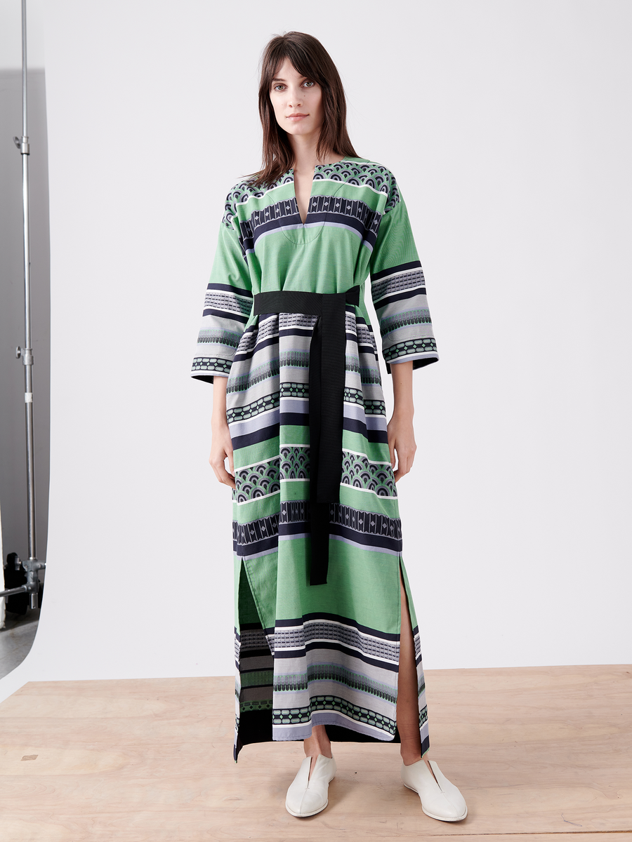 Someone is dressed in the MC Caftan by Zero + Maria Cornejo, a long robe with a green, black, and white pattern made from GOTS-certified cotton. It has wide sleeves and a black grosgrain tie belt, paired with light slip-on shoes on a plain wooden floor.