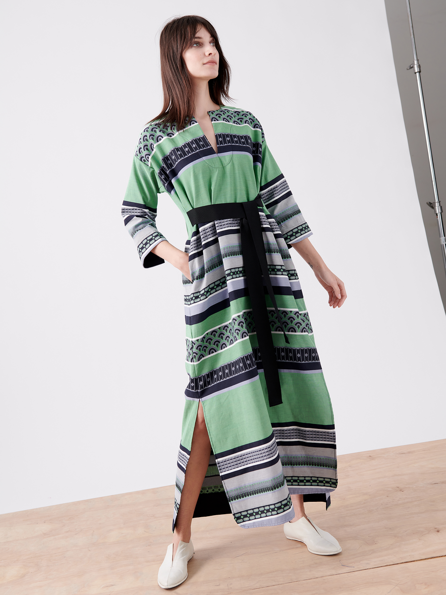 A person stands on a light wooden floor wearing the MC Caftan by Zero + Maria Cornejo, a long patterned green jacquard dress with black and white horizontal stripes. It features a black grosgrain tie belt and is paired with white slip-on shoes, as they gaze slightly to the side.