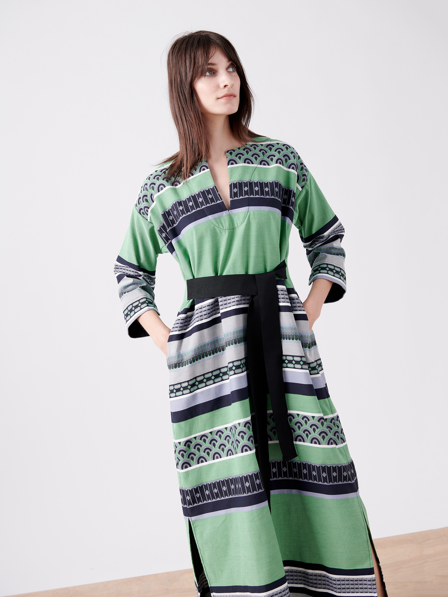 A woman poses in the MC Caftan by Zero + Maria Cornejo, a long green dress with geometric patterns and black accents. The GOTS certified cotton piece features a black belt. Her long dark hair frames her face against a plain light background, hands casually in pockets. Final sale item.