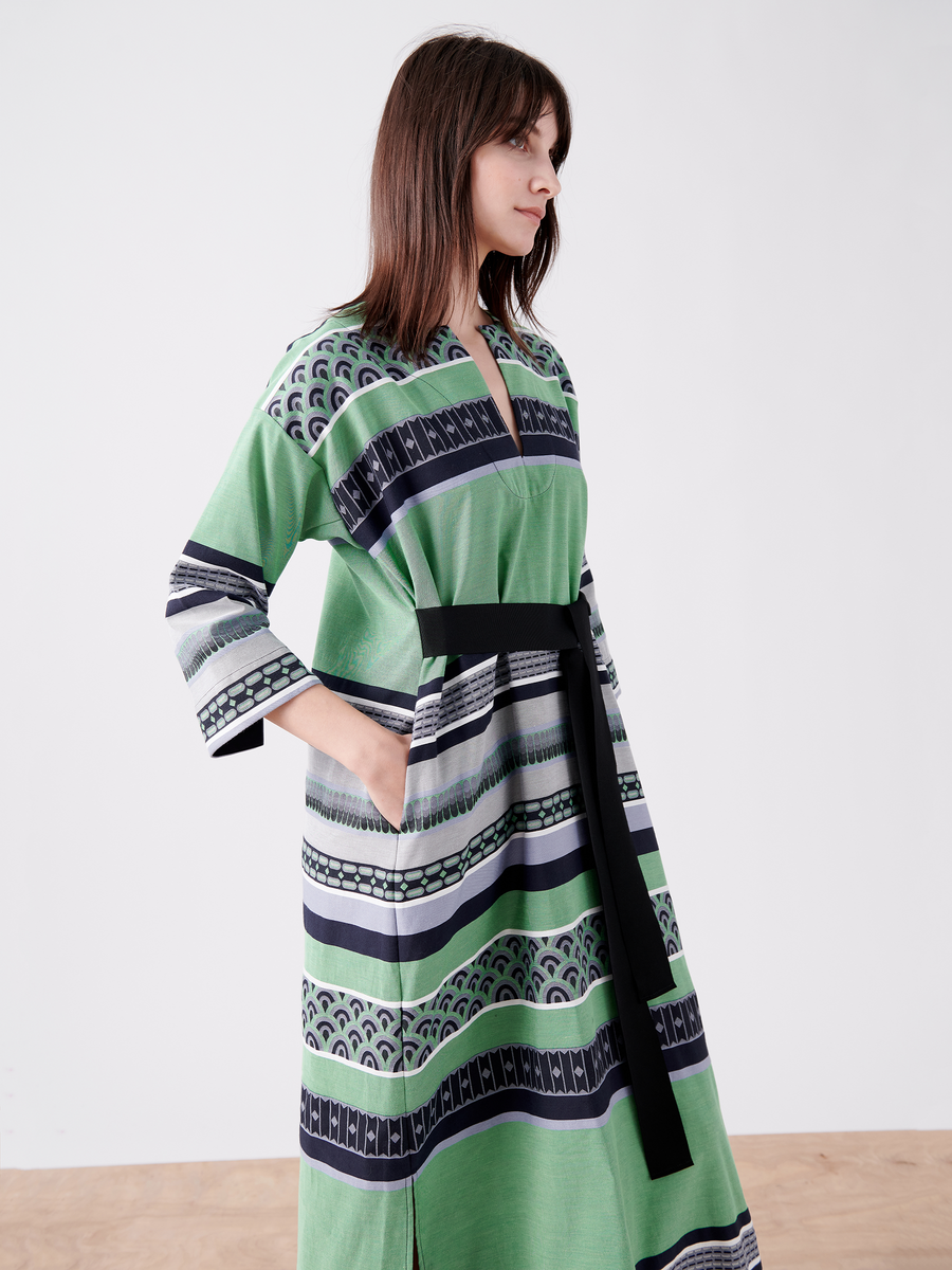 A person with long brown hair poses in profile against a white background, wearing the MC Caftan by Zero + Maria Cornejo. This green and black striped jacquard dress, on final sale, features a grosgrain tie belt, intricate geometric designs, long sleeves, and is made from GOTS-certified cotton.