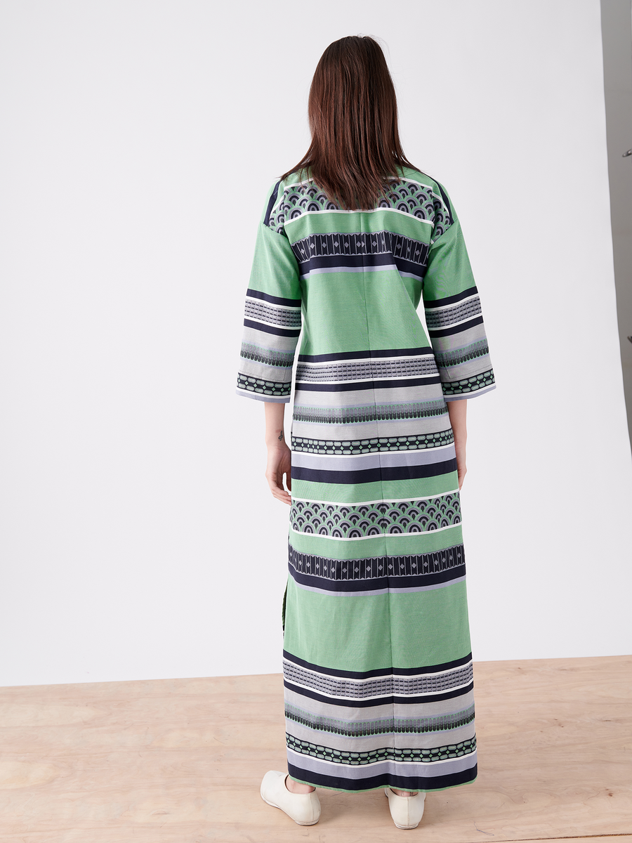 A person stands on a wooden floor with their back to the camera, wearing the MC Caftan from Zero + Maria Cornejo in green, blue, and black with a grosgrain tie belt and white shoes. The background is minimalist against a stark white wall.