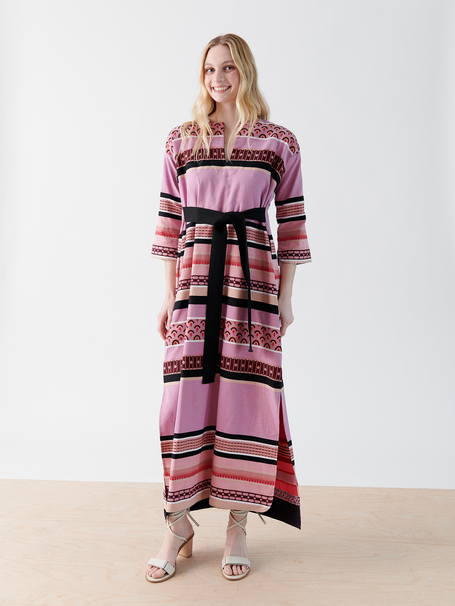 A person in the Zero + Maria Cornejo MC Caftan, a long pink patterned dress with a grosgrain tie belt and geometric designs in pink and black, stands smiling on a wooden floor. The individual has long, wavy hair.