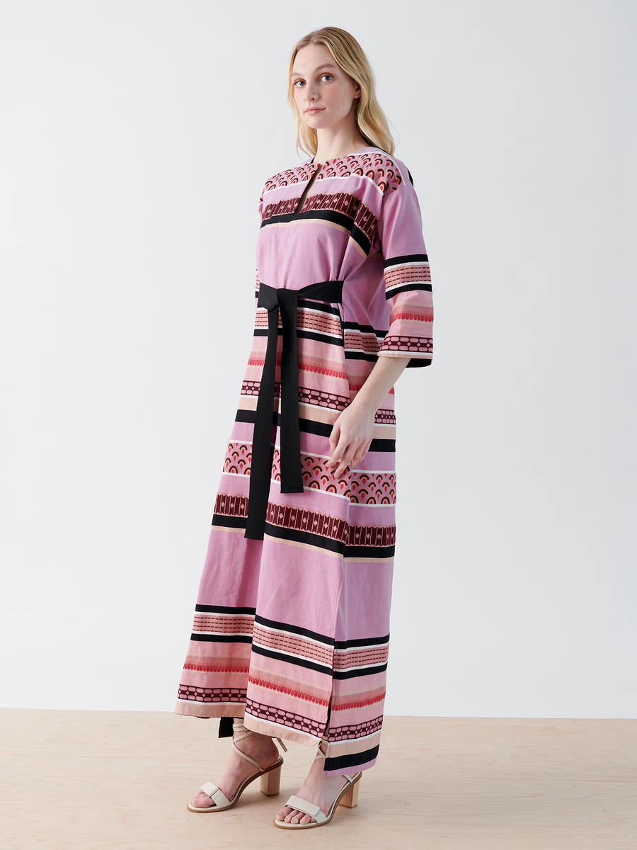 A person is modeling the MC Caftan from Zero + Maria Cornejo. It's a long pink dress with black and red geometric patterns featuring a grosgrain tie belt. They stand on a light wood floor, wearing beige open-toe heels against a plain white background.