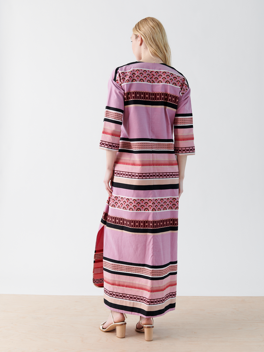 A person with long hair is wearing Zero + Maria Cornejo's MC Caftan—a long, patterned pink dress with red, black, and white horizontal stripes. This striped jacquard dress is made from GOTS-certified cotton and viewed from the back on a light wood surface.