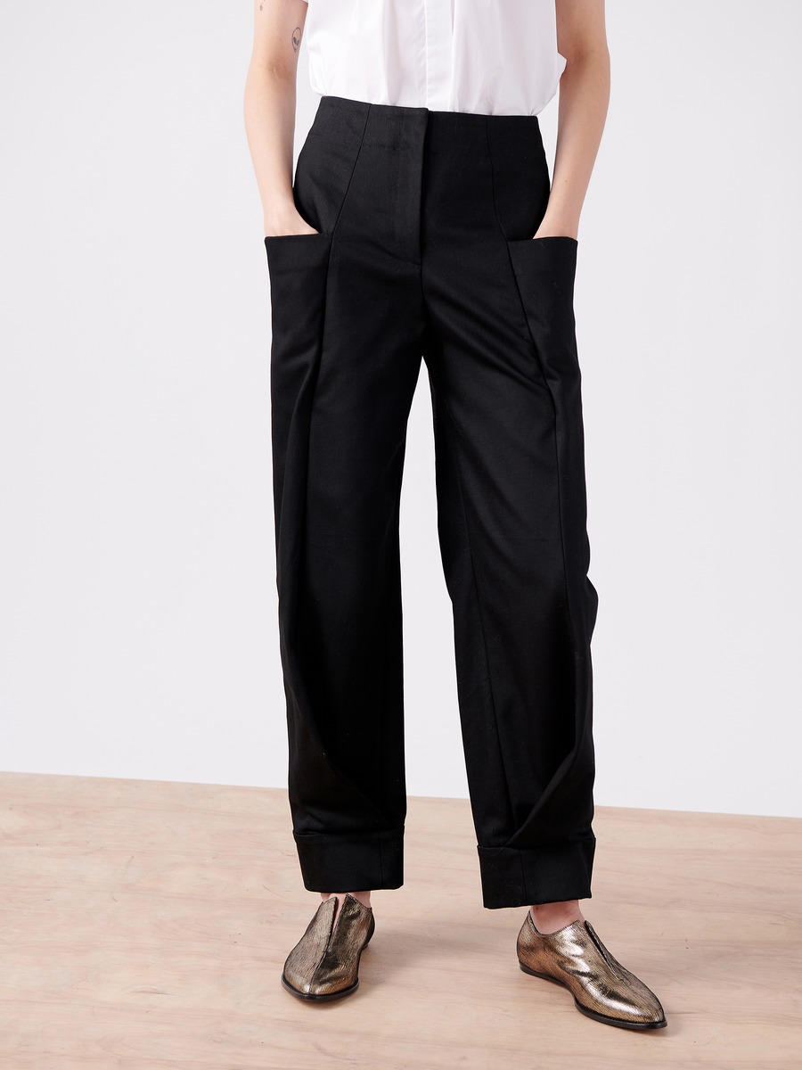 The individual sports a white 100% organic cotton shirt, tucked into black high-waisted trousers, with hands in pockets on a wooden floor. They complement the outfit with metallic pointed-toe shoes called Akeo Jodhpur from Zero + Maria Cornejo (Final Sale).