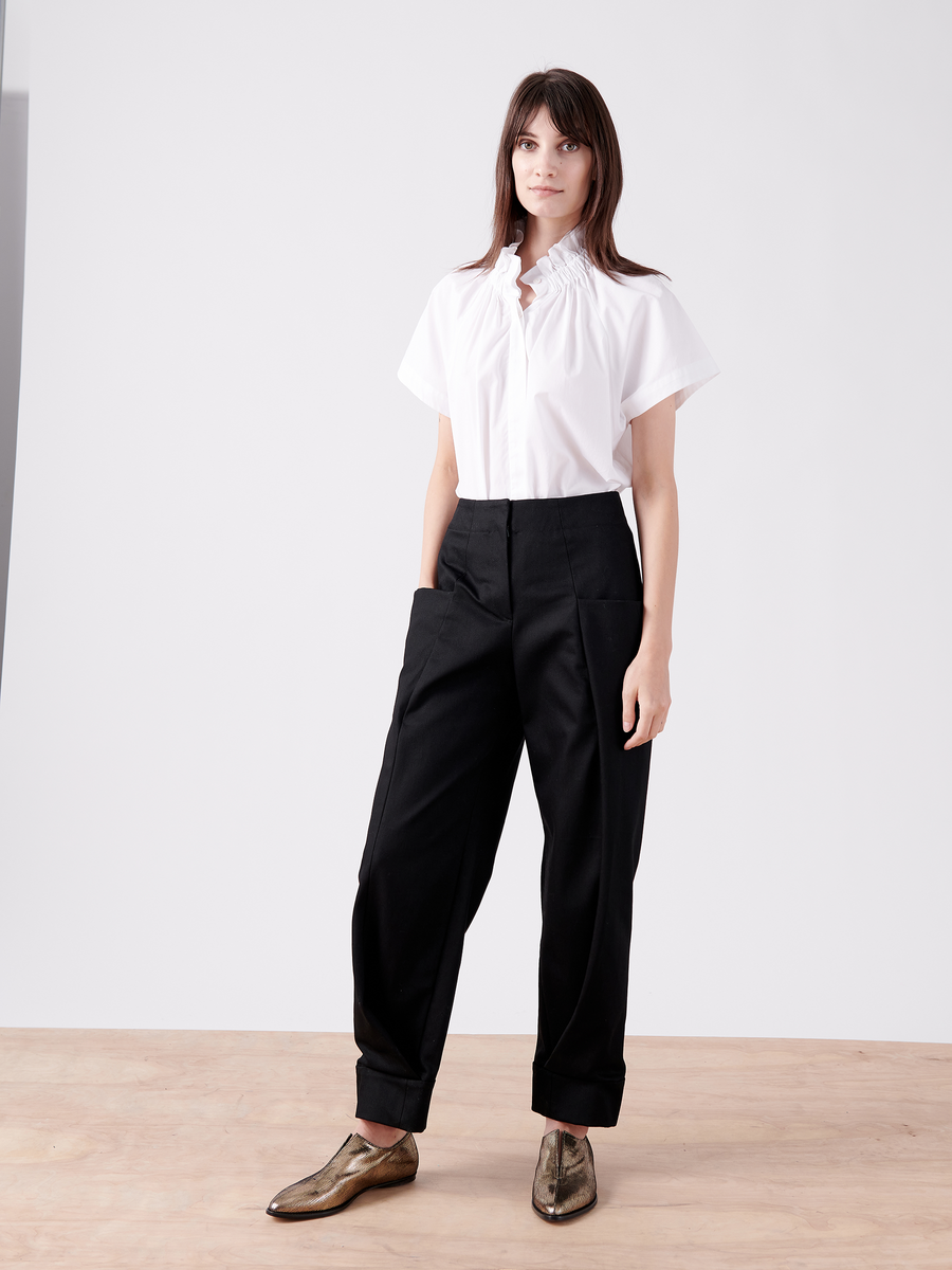 A person with long hair stands on a wooden floor against a white background, wearing a white short-sleeved blouse and shiny bronze Akeo Jodhpur by Zero + Maria Cornejo. Their look is completed with black 100% organic cotton trousers, as they gaze directly at the camera.