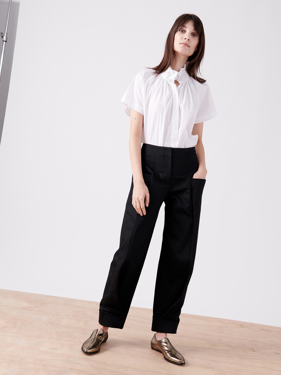 A person with straight brown hair stands on a wooden floor against a white background, wearing a 100% organic cotton white short-sleeve blouse and black trousers, with one hand in their pocket. Their look is completed by the shiny gold Akeo Jodhpur shoes from Zero + Maria Cornejo's final sale.