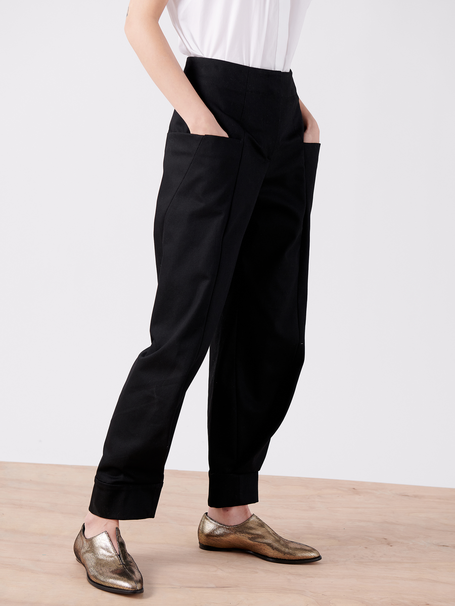 A person in high-waisted, Cradle 2 Cradle Certified black Akeo Jodhpur trousers from Zero + Maria Cornejo, hands in pockets, wears a white shirt and metallic bronze loafers on a wooden floor against a plain white background.