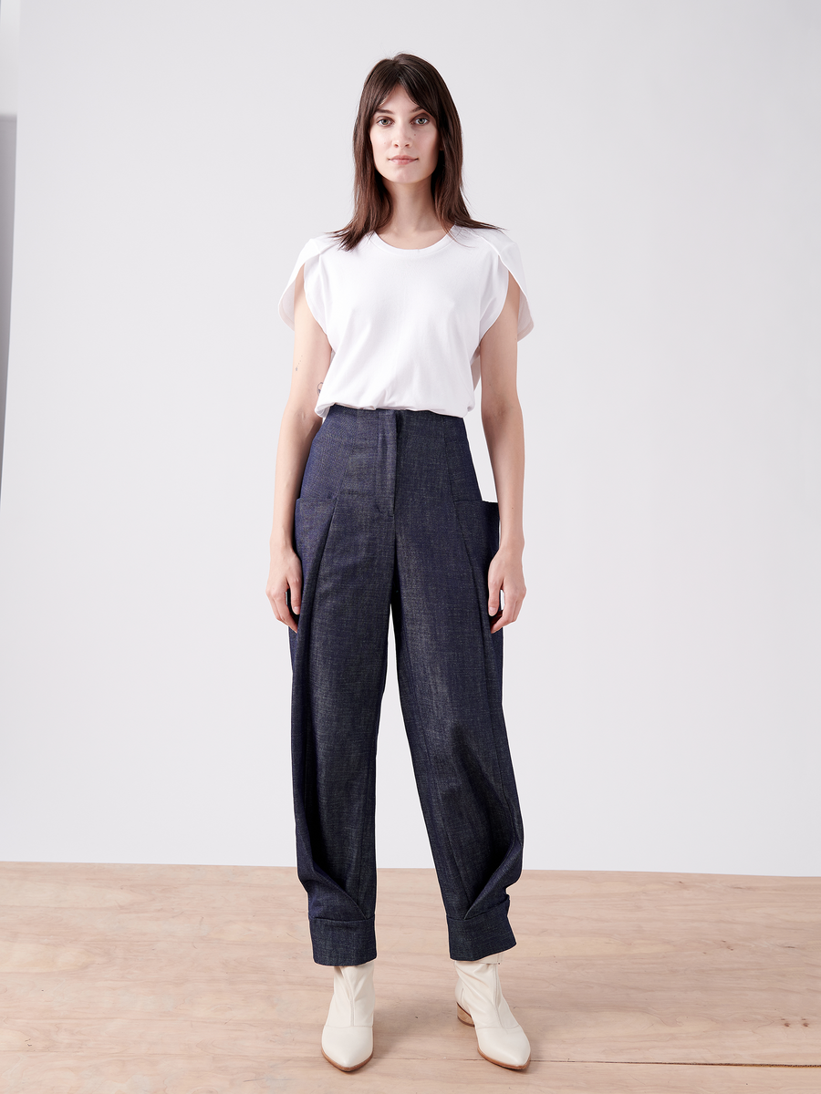 On a wooden floor with a plain backdrop, an individual wears a white t-shirt and Cradle 2 Cradle Certified™ organic denim pants. Their look is enhanced by Akeo Jodhpur boots from Zero + Maria Cornejo, while their shoulder-length hair frames a direct gaze at the camera.