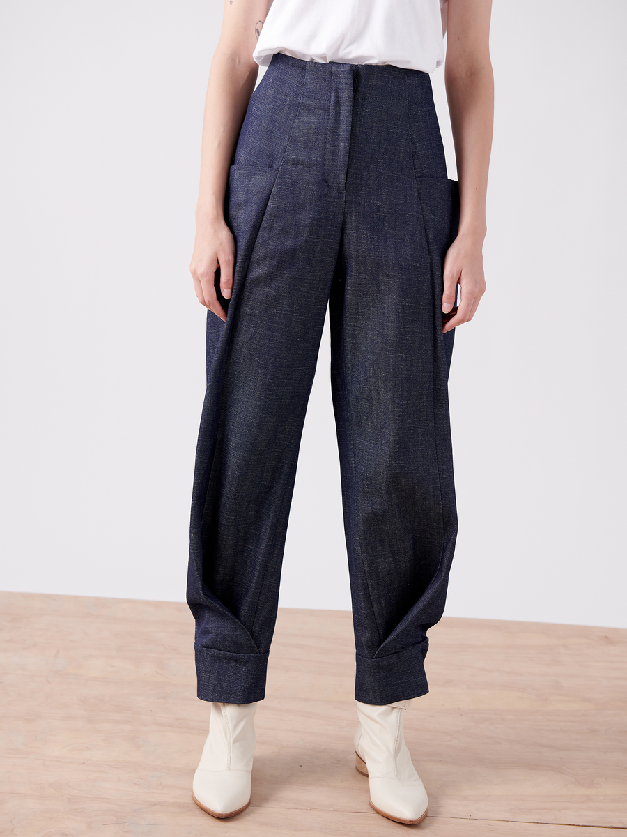 The individual is modeling the Akeo Jodhpur from Zero + Maria Cornejo, high-waisted wide-leg pants crafted from 100% organic denim with large pockets. They are paired with cream ankle boots on a wooden floor against a plain white backdrop.