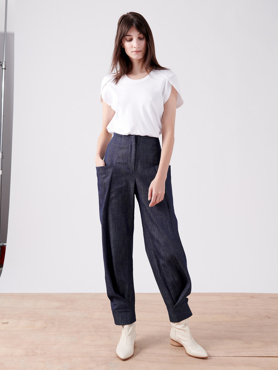 A person stands on a wooden floor in a white short-sleeved top, high-waisted loose-fitting dark pants made from organic denim, and light-colored ankle boots (Akeo Jodhpur - Final Sale by Zero + Maria Cornejo). The background is plain and minimalistic.