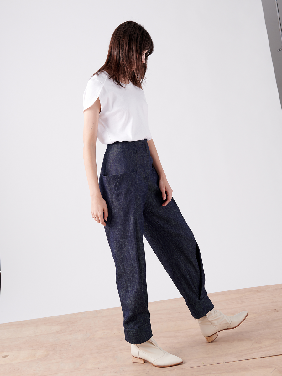 A person with shoulder-length brown hair wears a white T-shirt, high-waisted organic denim pants, and Akeo Jodhpur ankle boots by Zero + Maria Cornejo. They stand on a wooden floor against a plain white background, looking down.