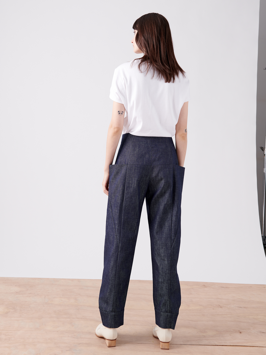 A person stands on a wooden floor, wearing a white T-shirt, Zero + Maria Cornejo Akeo Jodhpur 100% organic cotton high-waisted dark blue wide-leg pants, and light-colored shoes with their back to the camera against a plain white background.