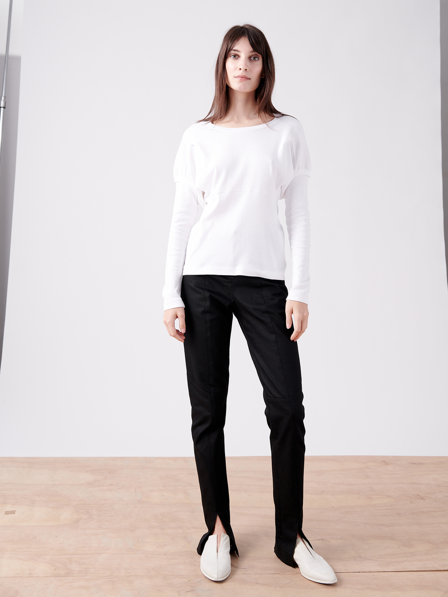 A person with long brown hair stands on a wooden floor against a plain backdrop, wearing a white organic cotton long-sleeve top, the Denim Tala Trouser (black with ankle slits) from Zero + Maria Cornejo, and white slip-on shoes.