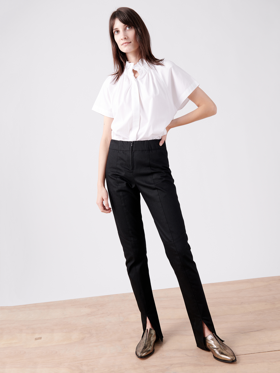 A person stands on a wooden floor wearing a white short-sleeve blouse and the Cradle 2 Cradle Certified Denim Tala Trouser by Zero + Maria Cornejo, featuring front slits. They have shoulder-length dark hair and shiny gold shoes, set against a plain white background.