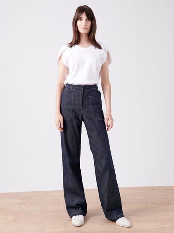 A woman stands on a wooden floor against a plain white backdrop. She is wearing a white T-shirt made from GOTS-certified cotton with slightly rolled sleeves and high-waisted, dark blue organic denim Eda Pant by Zero + Maria Cornejo. She has long brown hair and is wearing white shoes. She faces the camera with a neutral expression.