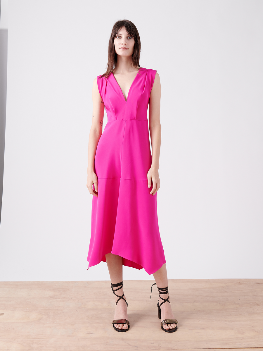 A woman poses gracefully on a wooden floor in the Joi Dress - Final Sale from Zero + Maria Cornejo. The sleeveless, bright pink cocktail dress features an asymmetrical hem. She complements it with black strappy heels, and her long dark hair cascades against a plain white background.