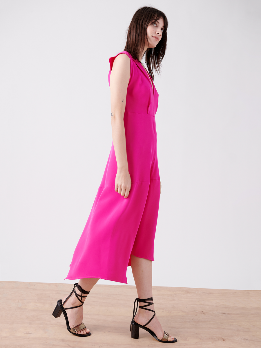 A woman with long dark hair wears the sleeveless knee-length Joi Dress in hot pink silk crepe by Zero + Maria Cornejo. She stands on a wooden floor in black strappy sandals against a plain white background, looking to the left.