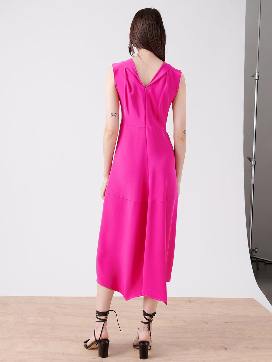 A person with long brown hair stands on a wooden floor, wearing the Zero + Maria Cornejo Joi Dress in vibrant pink silk crepe with a deep V back and irregular hem, paired with black strappy high-heeled sandals and small tattoos embellishing their arms.