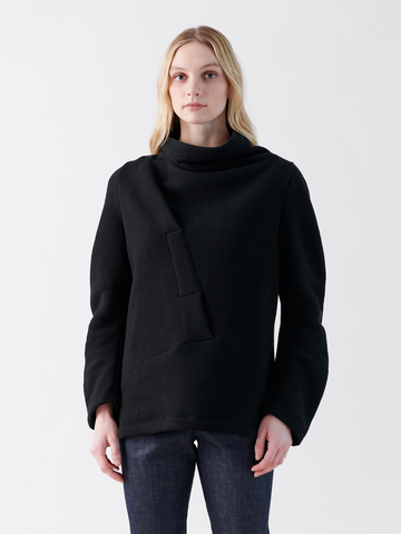 A person with long blonde hair wears Zero + Maria Cornejo's stylish Long-Sleeved Fold Top in black, showcasing an oversized silhouette and high neck. They stand against a plain white background, pairing the sweater with dark pants.