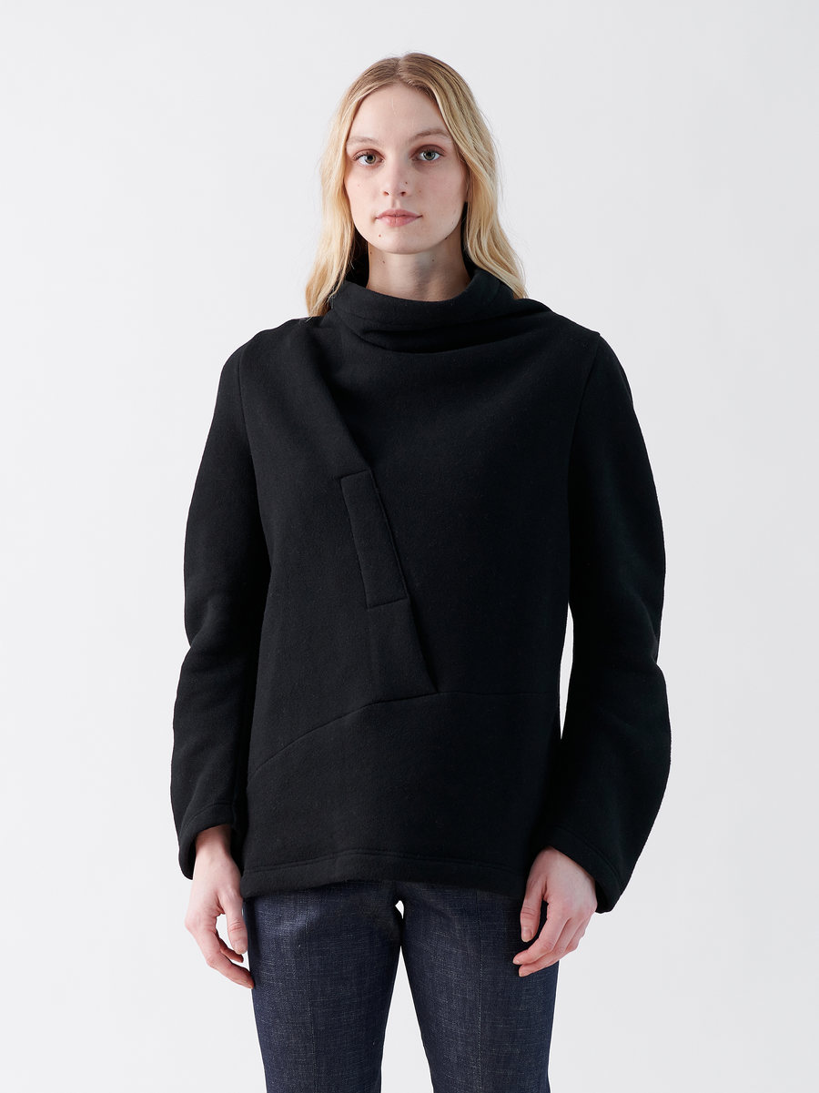 A person with long blonde hair wears Zero + Maria Cornejo's stylish Long-Sleeved Fold Top in black, showcasing an oversized silhouette and high neck. They stand against a plain white background, pairing the sweater with dark pants.