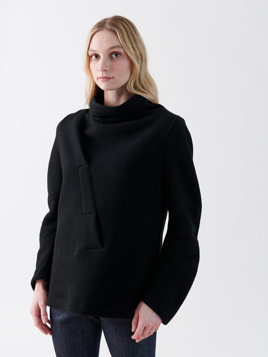 A person with long blonde hair wears the oversized Long-Sleeved Fold Top from Zero + Maria Cornejo—a loose-fitting black sweater made of biodegradable Lyocell—and dark jeans, standing against a plain white background.