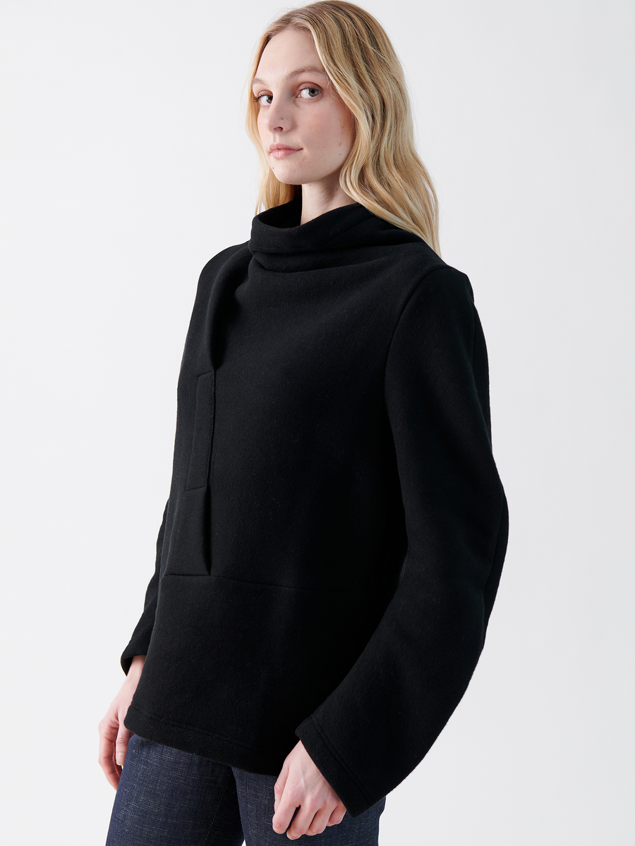 A person with long blonde hair wears an oversized Zero + Maria Cornejo black Long-Sleeved Fold Top and dark pants. They stand against a plain white background, looking slightly to the side with a neutral expression.