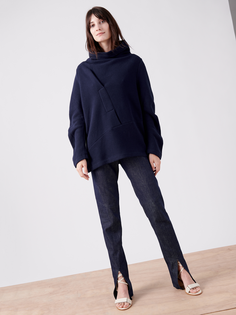 A woman poses in Zero + Maria Cornejo's oversized navy Long-Sleeved Fold Top made of biodegradable Lyocell, paired with dark front-slit jeans. Standing on light wooden flooring, she completes the stylish yet casual look with open-toe heels against a neutral background.