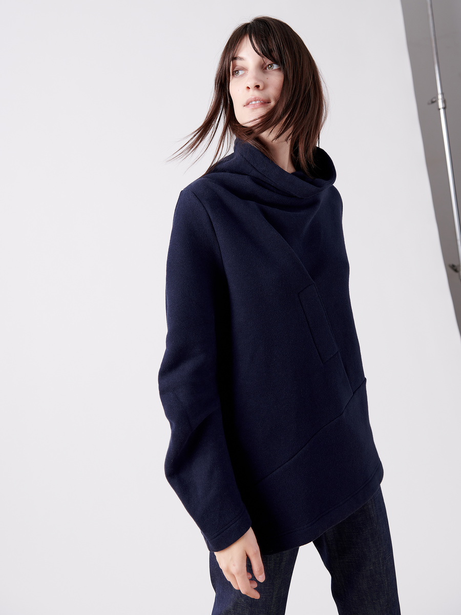 A person with shoulder-length hair wears a dark blue Zero + Maria Cornejo Long-Sleeved Fold Top from the final sale, featuring a high collar. They look to the side against a plain gray background.