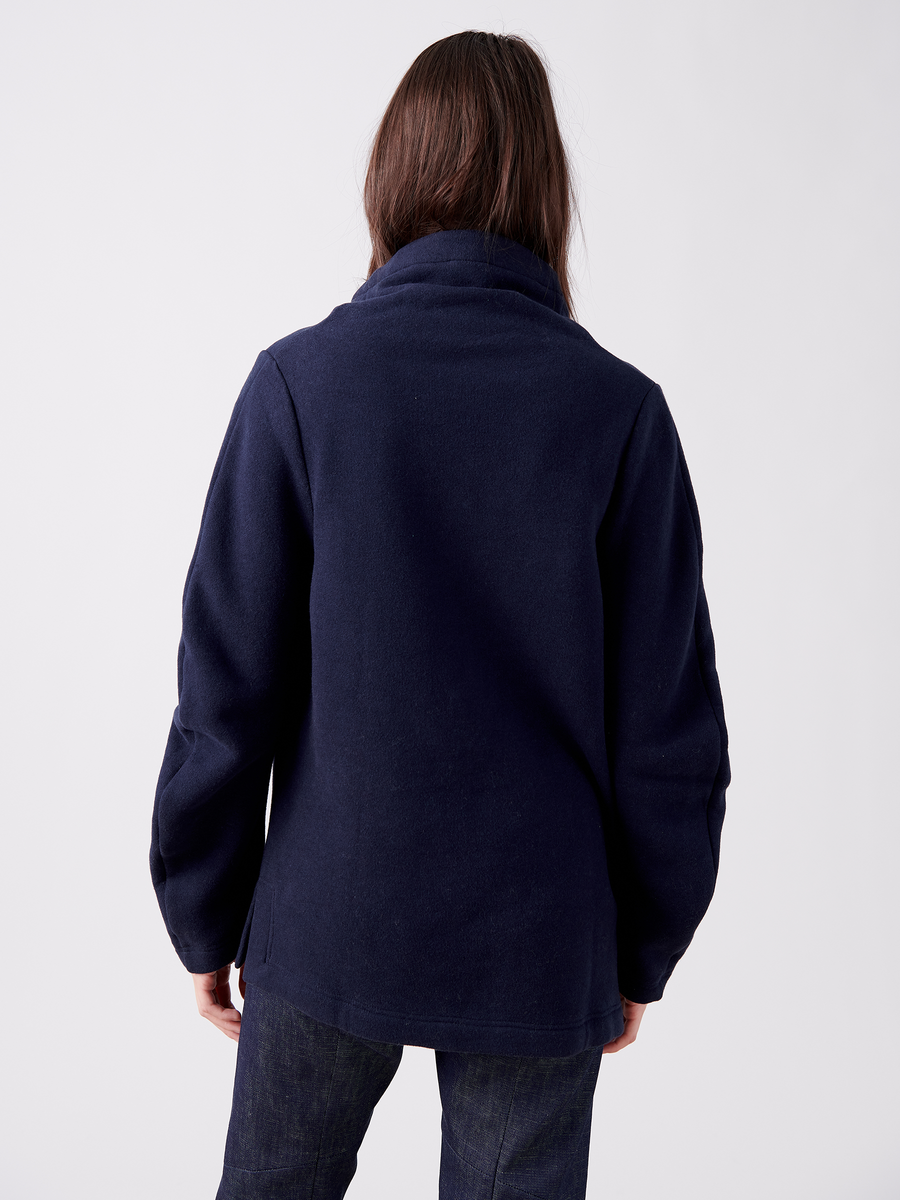 A person in an oversized silhouette wears a Zero + Maria Cornejo Long-Sleeved Fold Top paired with jeans, set against a light gray background. Their long dark hair contrasts with the sleek high-collared dark blue jacket's design.