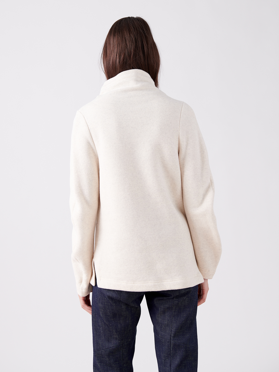 A person with long hair stands against a white background, wearing an oversized, cream-colored Long-Sleeved Fold Top by Zero + Maria Cornejo and dark blue jeans.