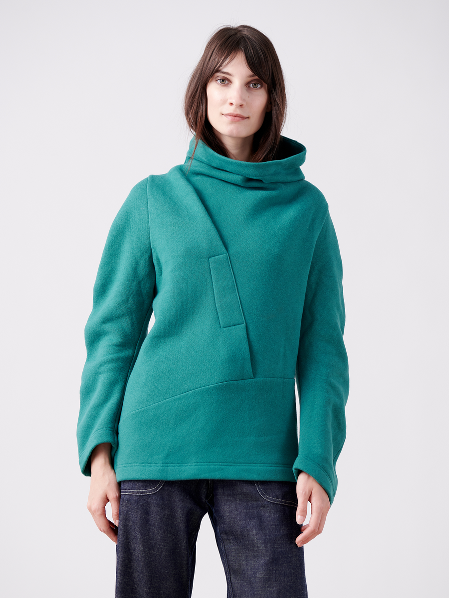 Against a white background, someone with long brown hair wears the Zero + Maria Cornejo Long-Sleeved Fold Top in an oversized teal style featuring an asymmetrical collar and geometric front design. Crafted from biodegradable Lyocell, they gaze directly at the camera.