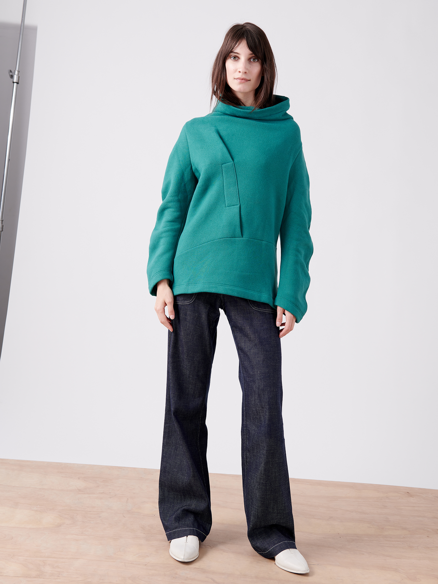 A person stands on a wooden floor wearing the Long-Sleeved Fold Top by Zero + Maria Cornejo, featuring a teal terrycloth material and asymmetrical collar design, dark wide-leg jeans, and white shoes. The background is a plain white wall with a softbox partially visible on the left.