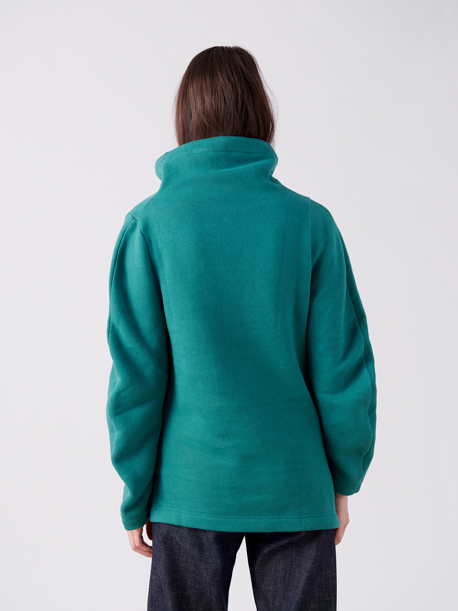 A person with long hair wears the Zero + Maria Cornejo Long-Sleeved Fold Top, an oversized teal high-collared jacket made from biodegradable Lyocell, and dark pants while facing away against a plain white background.