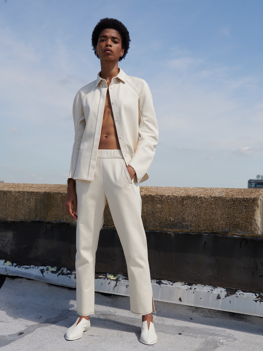 On a rooftop, someone stands confidently in an open white jacket and Denim Elli Pant by Zero + Maria Cornejo, revealing their torso against the serene blue sky. White shoes complete the minimalistic style, showcasing both fashion sense and eco-consciousness.