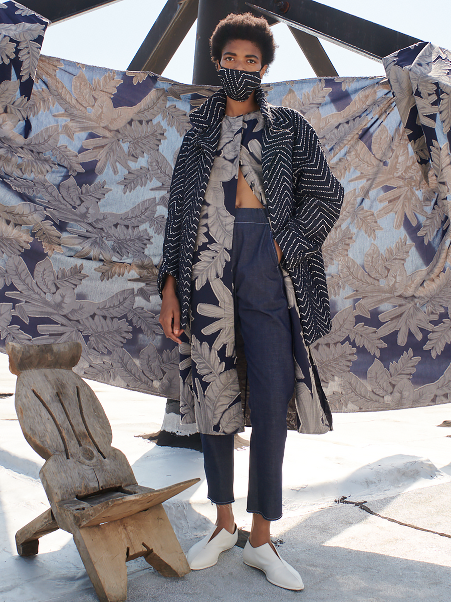 A person showcases an outfit from Zero + Maria Cornejo with Denim Elli Pant - Final Sale and a matching mask on a rooftop. The look features a long, layered coat and white shoes. Nearby, there’s a wooden chair and patterned fabric hanging in the background.