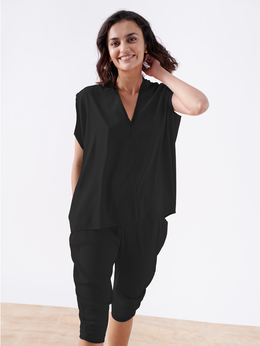 A person smiles while standing and touching their hair, dressed in an elegant loose-fitting black crop curve outfit from Zero + Maria Cornejo. The ensemble features a short-sleeved top and silk charmeuse pants that drape gracefully through the leg. The backdrop is a plain white wall with a light wooden floor.